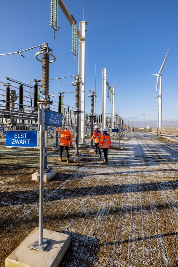 Drie mannen in oranje werkkleding staan bij een electriciteitscentrale. Op de voorgrond een bordje met 'elst zwart'