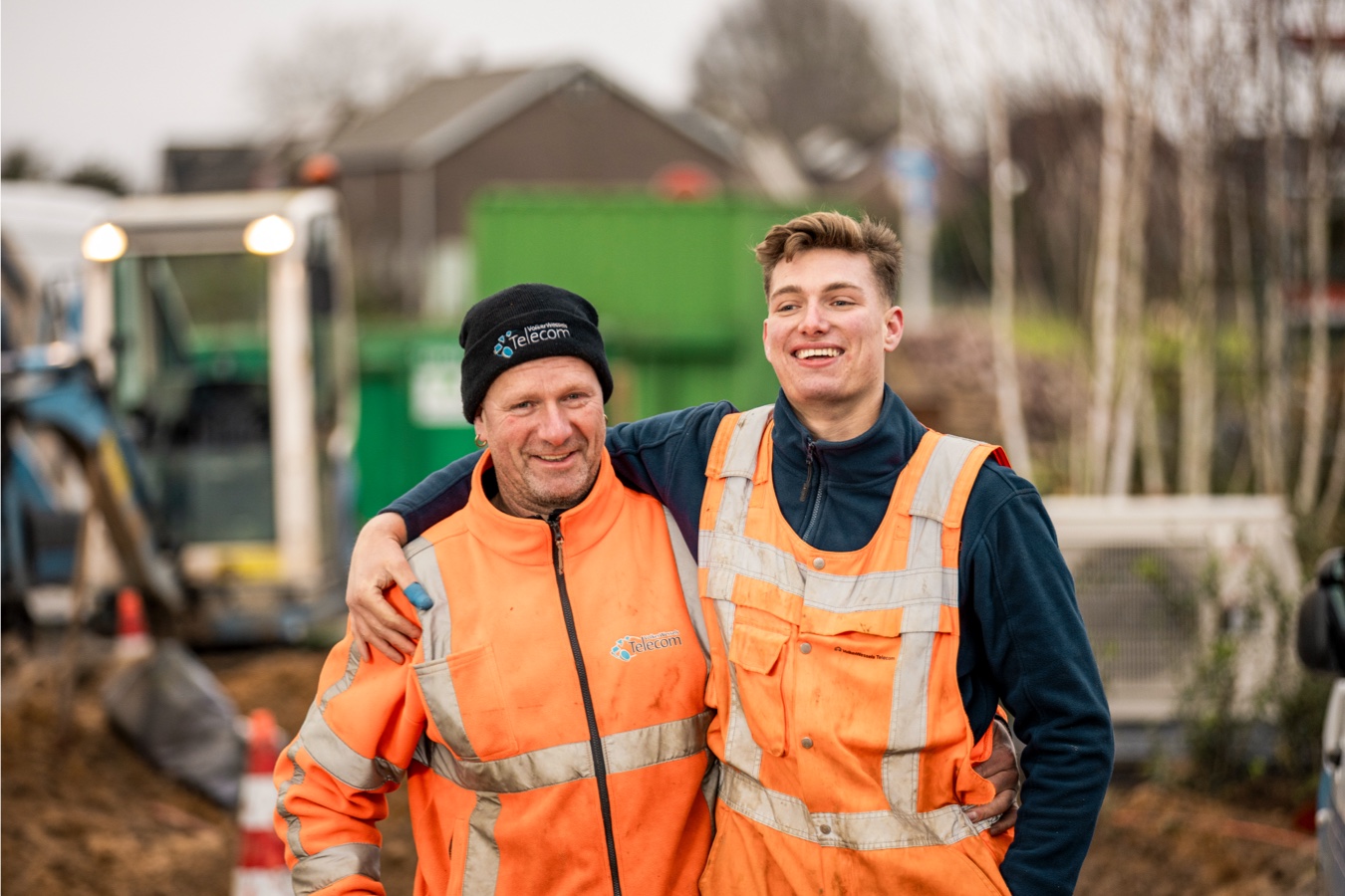 Twee lachende mannen staan op een bouwplaats en hebben een arm om elkaar heen geslagen.